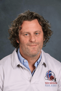 A man with curly hair smiles, wearing a light gray polo shirt with a school logo on it, against a dark background.