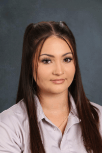 Una mujer joven con pelo largo castaño y un aro en la nariz, vestida con una camisa gris claro, posa sobre un fondo oscuro.